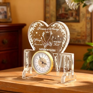 May include: A clear acrylic clock with a gold-tone clock face and the word "LOVE" in large letters. The clock has a heart-shaped top with the words "Forever & Always" and other text. The clock is on a wooden surface.