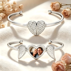 Könnte beinhalten: Ein silberfarbenes Armband mit einem herzförmigen Medaillon in der Mitte, das mit Engelsflügelmotiven und klaren Steinen verziert ist. Das Medaillon lässt sich öffnen und zeigt ein Foto einer älteren Frau und eines Mannes, während die Flügel ausklappen.
