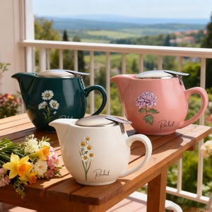 May include: Three ceramic teapots in teal, pink, and cream, each with a silver lid and a handle. The teal teapot features daisy illustrations, the pink one has a purple flower, and the cream one has daffodils. Each teapot has the name "Rachel" printed on it.