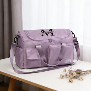 Peut inclure: Un sac de voyage lavande avec plusieurs poches et une bandoulière. Le sac porte le nom "Mohameo" imprimé en noir au-dessus d'un grand "M". Le sac est posé sur une table blanche.