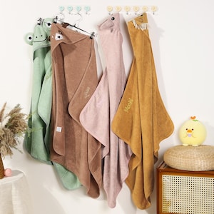 Pode incluir: Quatro toalhas com capuz para beb&ecirc;s em verde, marrom, rosa e dourado penduradas em uma parede branca. Cada toalha tem uma cara de animal no capuz: um sapo, um urso, uma raposa e um urso. As toalhas s&atilde;o macias e felpudas, perfeitas para a hora do banho.