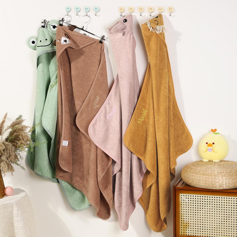 Pode incluir: Quatro toalhas com capuz para beb&ecirc;s em verde, marrom, rosa e dourado penduradas em uma parede branca. Cada toalha tem uma cara de animal no capuz: um sapo, um urso, uma raposa e um urso. As toalhas s&atilde;o macias e felpudas, perfeitas para a hora do banho.