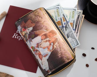 Puede incluir: Una cartera negra con cremallera y cremallera y tirador dorados. La cartera presenta una impresión fotográfica familiar. Varios billetes de cien dólares son visibles en el interior. La cartera está sobre una superficie blanca.
