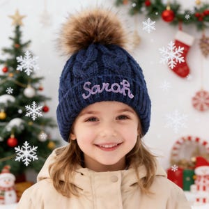Op de afbeelding: Een marineblauwe kabelgebreide beanie met een bruine imitatiebont pompon. De naam "Sarah" is in lavendel op de voorkant geborduurd. De achtergrond bevat een kerstboom en sneeuwvlokdecoraties.