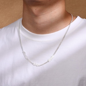 Op de afbeelding: Zilverkleurige ketting met drie naamhangers: "Amelie", "Logan" en "Maria". De ketting wordt om de nek gedragen en hangt over een wit T-shirt. De namen zijn in een cursief lettertype.