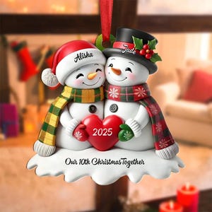 May include: A snowman Christmas ornament featuring two snowmen embracing, holding a red heart with "2025" on it. One snowman wears a red hat and scarf, the other a black hat and scarf. The ornament reads "Alisha" and "Jade".