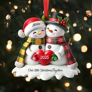 May include: A Christmas ornament featuring two snowmen embracing. One snowman wears a red Santa hat with the name "Alisha", and the other wears a black top hat with the name "Jade". They hold a red heart with "2025" on it. The ornament reads "Our 10th Christmas Together".