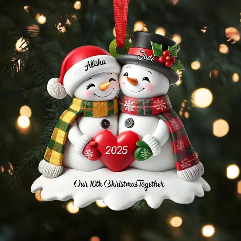 May include: A Christmas ornament featuring two snowmen embracing. One snowman wears a red Santa hat with the name "Alisha", and the other wears a black top hat with the name "Jade". They hold a red heart with "2025" on it. The ornament reads "Our 10th Christmas Together".