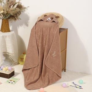 Pode incluir: Uma toalha de banho com capuz castanha para beb&eacute; com um desenho de rosto de urso no capuz. A toalha est&aacute; pendurada sobre um arm&aacute;rio de madeira. A palavra "Jesus" &eacute; bordada na toalha. A toalha &eacute; feita de material macio.