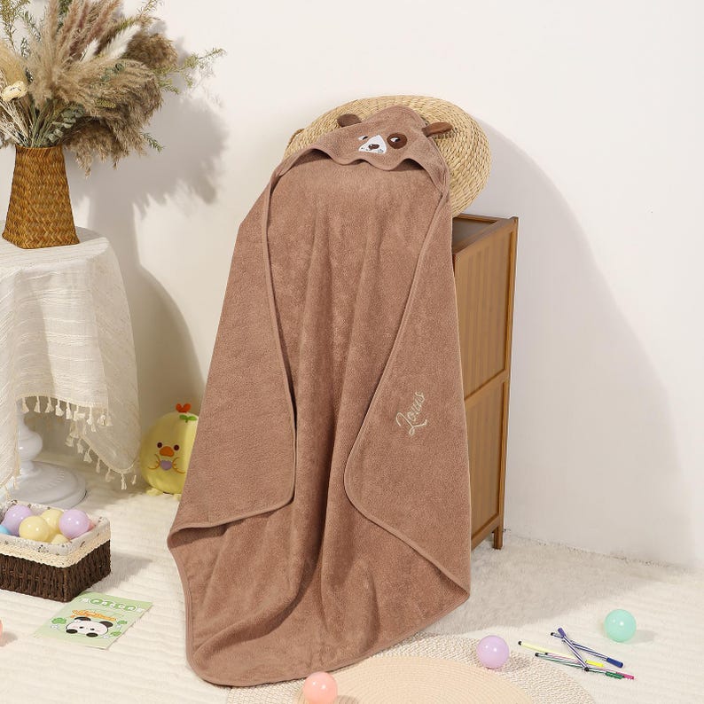 Pode incluir: Uma toalha de banho com capuz castanha para beb&eacute; com um desenho de rosto de urso no capuz. A toalha est&aacute; pendurada sobre um arm&aacute;rio de madeira. A palavra "Jesus" &eacute; bordada na toalha. A toalha &eacute; feita de material macio.