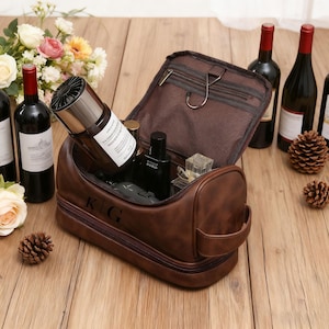 Puede incluir: Un neceser de cuero marrón con las iniciales "KG" está abierto, revelando varios productos de aseo. La bolsa tiene una cremallera y un asa. Botellas de vino y flores en el fondo.