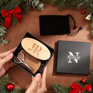 May include: A black gift box containing a wooden beard brush, comb, and scissors. The brush is engraved with "JR Jeremiah". A black pouch and a separate black box with "NICHOLAS" printed on it are also present. The scene is decorated with Christmas ornaments.