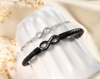 May include: Two infinity symbol bracelets. One is silver with clear stones and the other is black leather with a black infinity symbol. The bracelets are displayed on a white, curved surface. The silver bracelet has the name "Sarah David" engraved on it. The black bracelet has the name "Mireyal Encayi" engraved on it.