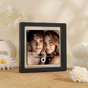 May include: A black square frame with a photo of two people, with the text "Because You Live" and "Jesse McCartney". The frame is on a wooden surface, with white flowers and a decorative vase in the background.