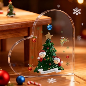 Könnte beinhalten: Ein herzförmiger, klarer Weihnachtsschmuck mit einem grünen Weihnachtsbaum und einem goldenen Stern. Zwei Figuren mit Weihnachtsmützen flankieren den Baum. Der Schmuck trägt den Text "Merry Christmas" auf einem Banner.