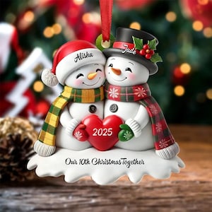 May include: A Christmas ornament featuring two snowmen embracing, holding a red heart with "2025" on it. One snowman wears a red hat and scarf, the other a black top hat and scarf. The ornament reads "Our 10th Christmas Together".