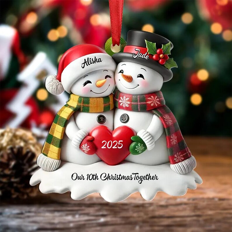 May include: A Christmas ornament featuring two snowmen embracing, holding a red heart with "2025" on it. One snowman wears a red hat and scarf, the other a black top hat and scarf. The ornament reads "Our 10th Christmas Together".