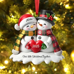 May include: A Christmas ornament featuring two snowmen embracing. One snowman wears a red Santa hat and a green and gold scarf, the other a black top hat and a red and green scarf. They hold a red heart with "2025" on it. The ornament reads "Our 10th Christmas Together."