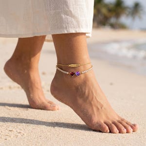 Peut inclure: Un ensemble de deux bracelets de cheville dorés. La chaîne supérieure comporte une fine barre dorée avec un texte gravé et un symbole de cœur. La chaîne inférieure est un bracelet de cheville de style tennis avec une pierre rouge et une violette.