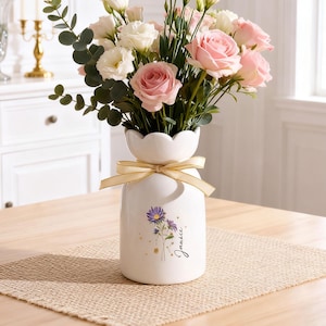 Puede incluir: Un jarrón de cerámica blanco con borde festoneado, atado con una cinta dorada, contiene un ramo de rosas rosas y blancas y vegetación. El jarrón está decorado con un diseño floral y el nombre "Janice".