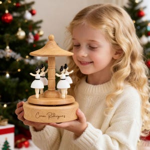 May include: A wooden carousel music box featuring four ballerina figurines in white dresses and black hair. The music box has a light brown finish and a decorative top. The base of the music box has the inscription "Cesar Rodrigues".