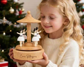 May include: A wooden carousel music box featuring four ballerina figurines in white dresses and black hair. The music box has a light brown finish and a decorative top. The base of the music box has the inscription "Cesar Rodrigues".