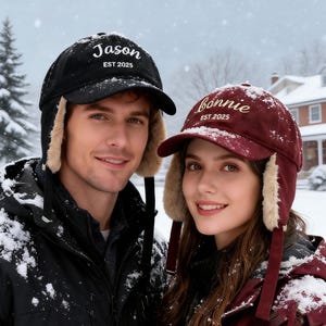 Peut inclure: Deux bonnets d'hiver avec cache-oreilles. L'un est noir avec "Jason EST 2025" brodé en blanc. L'autre est bordeaux avec "Bonnie EST 2025" brodé en or. Les deux chapeaux ont une doublure en fausse fourrure et sont portés dans un environnement enneigé.