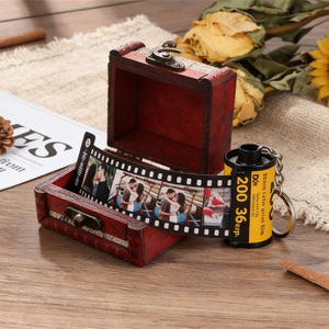 May include: A small, open, red wooden chest with a metal clasp holds a strip of photos. A yellow film canister keychain with black text is next to the chest. The film strip displays several photos of a couple.