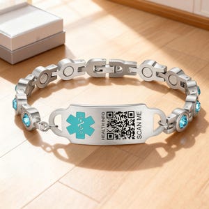 以下が含まれることがあります： ターコイズブルーの医療シンボル、QRコード、および「HEALTH INFO SCAN ME」のテキストが特徴の、シルバーの医療IDブレスレット。ブレスレットには、青い宝石のアクセントと留め金が付いています。