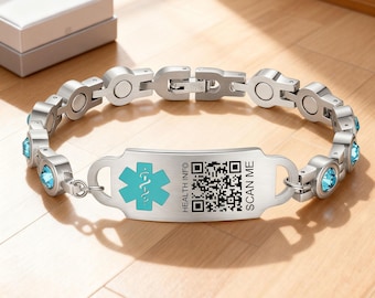 Puede incluir: Una pulsera de identificación médica plateada con una placa central con un símbolo médico turquesa, un código QR y el texto "HEALTH INFO SCAN ME". La pulsera tiene detalles de piedras preciosas azules y un cierre.