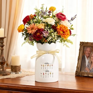 Puede incluir: Un jarrón de cerámica blanco con la parte superior festoneada, lleno de un vibrante arreglo de flores coloridas. El jarrón tiene una cinta dorada atada alrededor del cuello y presenta las palabras "Mum" y "Nan" con ilustraciones de flores. Una foto enmarcada está a la derecha.