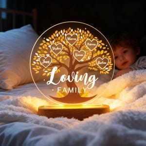 Pode incluir: Uma luz noturna personalizada com árvore genealógica. O disco de acrílico transparente apresenta um desenho de árvore com nomes em folhas em forma de coração. A base é de madeira. As palavras "Loving Family" estão escritas em itálico.