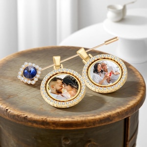 May include: Gold-colored personalized photo pins with a round pendant. The pendants are adorned with small, clear crystals around the edges. One pin features a blue gemstone surrounded by crystals. The pins display photos of couples.