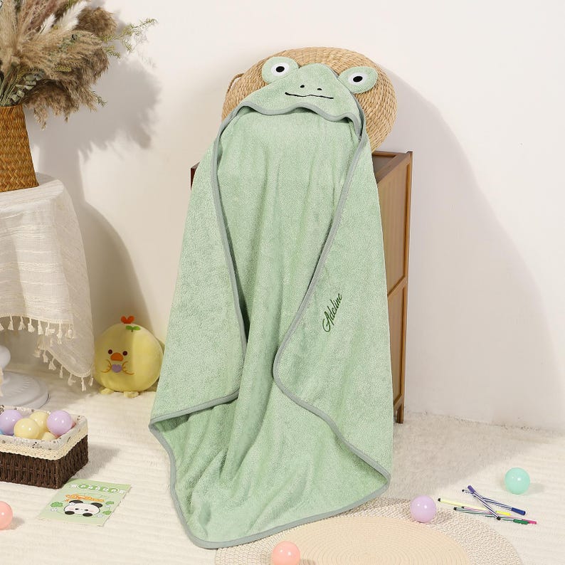 Pode incluir: Uma toalha de banho com capuz verde claro para beb&eacute; com um desenho de rosto de r&atilde;. A toalha tem uma borda cinzenta e o nome "Adeline" bordado na frente. A toalha est&aacute; sobre um cesto de vime.