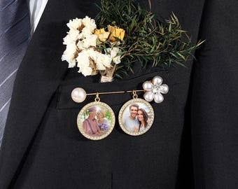 Puede incluir: Un broche dorado con dos colgantes circulares para fotos enmarcados con pequeños cristales transparentes, una perla blanca y una flor blanca de seis pétalos con un cristal central, prendido en una chaqueta de traje negra con follaje.