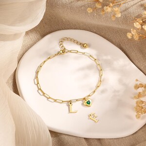 May include: Gold-colored bracelet with a chain link design. The bracelet features a heart-shaped charm with a green gemstone and the letter "L". Additional letter charms are also present. The bracelet is displayed on a white, curved surface.