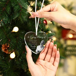 Può includere: Un ornamento di vetro trasparente a forma di cuore con la scritta "In Loving Memory of" e una foto di un uomo. L'ornamento è sospeso da un nastro bianco su uno sfondo di albero di Natale. Il testo include il nome "Grandpa" e le date.