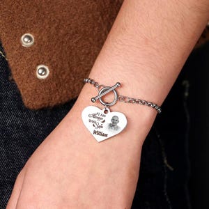 Può includere: Un braccialetto argentato con un ciondolo a forma di cuore. Il ciondolo presenta una foto e la scritta "I Am Always With You William". Il braccialetto ha una chiusura a levetta e viene indossato al polso.