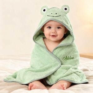 Puede incluir: Una toalla con capucha para bebé de color verde claro con un diseño de cara de rana. La toalla tiene un ribete gris y el nombre "Adeline" bordado en la parte delantera. El bebé está envuelto en la toalla y sentado en una cama.
