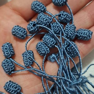 Op de afbeelding: Close-up van verschillende blauwe, geweven, kraalachtige versieringen op dunne blauwe draden. De kleine, ovaalvormige kralen zijn samengeklonterd en de draden worden in een hand gehouden. Deze kunnen worden gebruikt voor knutselen of het verfraaien van kleding.