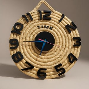 May include: Round woven wall clock with black numerals and hands. The clock face is made of natural-colored woven material with a black center. The word "TIME" is printed on the clock face. The hands are blue and pink.