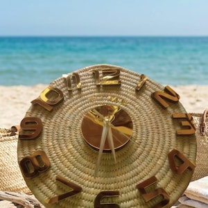 May include: A round, woven clock with gold-colored numerals and hands. The clock face is made of a natural, light brown material. The clock is displayed on a sandy beach with the ocean in the background.