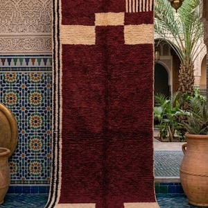 Peut inclure: Un tapis rectangulaire en laine avec un centre bordeaux profond et des accents géométriques couleur crème. Les extrémités présentent un motif à damier noir et blanc ainsi que des rayures verticales, terminées par une frange couleur crème.