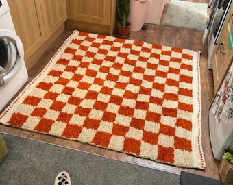 May include: A rectangular area rug featuring a repeating checkerboard pattern in vibrant orange and off-white. The rug has a thick, textured pile and is finished with a decorative stitched border along the edges, creating a distinct grid design.