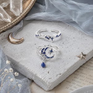 Può includere: Due anelli d'argento con disegni celesti. Uno presenta una luna crescente con una pietra blu pendente, stelle e dettagli blu. L'altro anello ha una stella e dettagli blu. Entrambi gli anelli sono su una superficie strutturata.