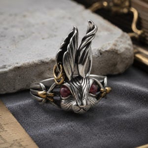 May include: A silver-toned metal ring featuring a sculpted rabbit head with prominent ears, deep red circular eyes, and small gold-toned accents including a tiny ring attached to one ear and arrow-like details on the sides of the band.