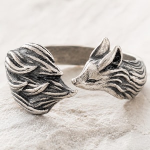 May include: A silver fox ring. The ring features a detailed fox head and tail design, with textured fur detailing. The ring's band is open, and the fox's head and tail meet at the ends. The ring is set against a white surface.