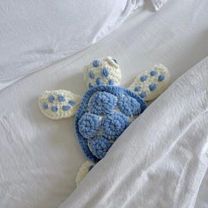 Peut inclure: Une peluche tortue de mer faite au crochet, composée d'un corps couleur crème avec des taches bleu clair sur les nageoires et la tête. La carapace est formée de segments de laine texturés et arrondis, alternant le bleu clair et le crème.
