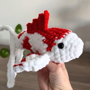 Könnte beinhalten: Ein gehäkeltes Plüschfisch-Spielzeug aus weichem, flauschigem Garn mit einem leuchtend roten und weißen Farbmuster. Der Fisch hat eine markante Rückenflosse, einen abgerundeten Kopf mit einem sichtbaren schwarzen Kunststoffauge und lange Schwanzflossen.