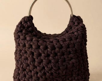 Puede incluir: Un bolso tote marrón oscuro tejido a mano con un asa grande de metal plateado. El bolso tiene forma cuadrada y un aspecto texturizado y tejido. El asa es un gran anillo semicircular.